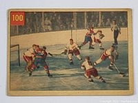 Front image of vintage 1954-55 Parkhurst hockey card numbered 100 showing hockey players around the net, including goalie Terry Sawchuk defending goal against Bernie Geoffrion's attempt.