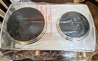 Photo of the dual burner stove wrapped in plastic, showing two black round burners on white base.