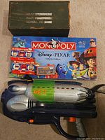 Top-down photo showing Disney Pixar Monopoly board game box, a toy rocket launcher gun, and a green metal box closed with name, rank, and serial number space on top