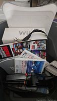 Photo of lot items inside a plastic bin including Wii console box, Rapala fishing rod accessory, game cases, and Wii Fit board.