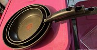 Photo of three black non-stick frying pans stacked, showing their handles and cooking surfaces with circular patterns indicating non-stick coating.