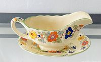 Side view of the ceramic gravy boat with attached handle and spout alongside the matching oval plate showing colorful floral pattern.