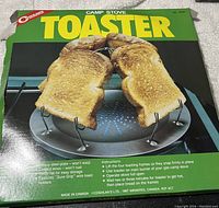 Coghlan's camp stove toaster in original packaging showing toasted bread slices on wire holders over burner plate.