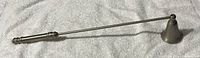 Metal candle snuffer with elongated handle and bell-shaped snuffer end displayed on a fabric surface.
