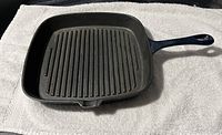 Square black cast iron grill frying pan with ridged surface and handle lying on white cloth.