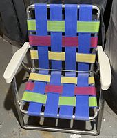 Photo of child-size folding camp chair showing colorful woven webbing (blue, green, red, yellow) on aluminum frame with white plastic armrests.