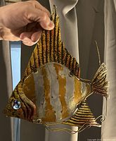 Metal fish wall decor held by hand to show design details including striped yellow and brown pattern and curled wire details for mouth and fins.