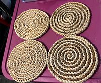 Four woven round potholders arranged on a pink surface, showing spiral braided pattern in light beige and dark natural tones.