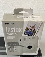 Box packaging of FujiFilm Instax Mini 9 instant camera in smoky white color, showing the front of the camera, branding, and some features like selfie mirror and brightness settings.