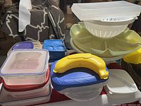 Photo showing assorted plastic food containers stacked with lids, a banana-shaped yellow container, and white salad spinner/colander bowl setup.