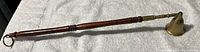 Wooden handle candle snuffer with brass bell on a fabric background, showing full length with a ring at the handle end