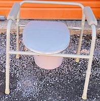 Portable toilet with metal frame, padded handles, and toilet seat lid shown outdoors on gravel surface.