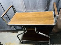 Photo of retro cart showing wood tray surface framed in chrome metal with wooden rail handles and chromed geometric legs.