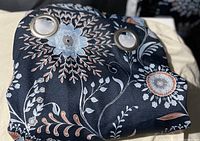 Close-up photo of folded navy blue curtain fabric showing floral pattern with large flowers in light blue, beige, and white. Two silver metal grommets are visible for hanging.