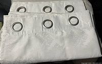 Photo shows two white curtains with an embossed damask pattern and silver metal grommets aligned at the top for hanging.