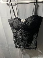Black lace cami top hanging on a white hanger against a curtain background. Top shows floral and mesh lace pattern with built in cups and adjustable spaghetti straps.