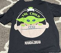 Front of black T-shirt showing The Child (Grogu) in hover pram with frog and text 'THE CHILD' and 'THE MANDALORIAN'.