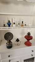 Full view showing all eight oil lamps arranged on shelves and surface with various glass colors and styles.