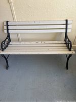 Front view of wood and metal park bench showing 3 horizontal white wooden slats for the backrest and 5 slats for the seat with black metal legs and armrests.