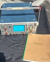 Angle view of oscilloscope unit with blue perforated metal housing, grey front panel controls, and the manual laid beside it on black surface.