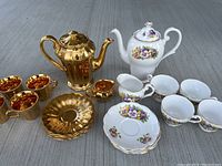 Photo showing both tea sets together with 4 gold cups, gold teapot, gold cream pitcher, white floral teapot, white cream pitcher, 4 white floral cups, and floral saucers.