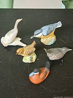 Five assorted porcelain and ceramic bird figurines arranged on a black surface, showing various colors and bases.