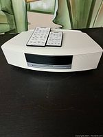 White Bose Wave Music System main unit on dark surface with two remotes placed on top