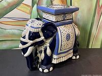 Front and side angle view of the ceramic elephant plant stand, showing blue and white detailed embossed patterns and tassels