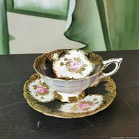 Angled view showing the teacup resting on the saucer, highlighting the gold ornate rim and pink rose floral design on black background.