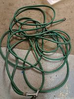 Photo showing coiled green garden hose on concrete floor, with visible metal connector end.