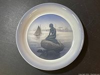 Collector's plate with image of The Little Mermaid statue on a rock by the harbor, showing sailboat and industrial background.