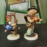 Front view of both Hummel figurines showing girl in tree branch and boy playing violin.