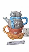 Front view of the cat-themed teapot set stacked on the teacup with curled orange cat figurine alongside and product tag visible