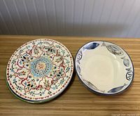 Two stacks of ceramic plates on a wooden surface. One stack has colorful floral and geometric patterns, the other stack has blue seafood-themed plates with scalloped edges.