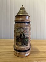 Front and side view of vintage German beer stein showing colorful Munich Olympiastadt image with Olympic rings and metal lid
