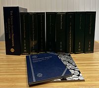 Photo of multiple Littleton coin albums on a wooden surface, mostly dark green and blue hardbound albums with gold lettering on spine showing different coin series and years.