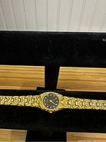 Full front view of gold-tone quartz watch displayed on black velvet horizontal holder showing band detail and black dial face.