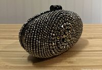 Oval pewter beaded clutch displayed on wooden surface showing front angled view, highlighting beaded details and black metal clasp.