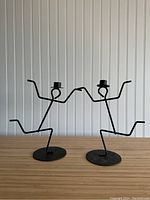 Pair of black metal stick figure candle holders standing side by side with arms and legs outstretched, placed on wood surface against white beadboard background.