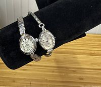 Two vintage Waltham ladies wristwatches displayed on black velvet watch stands. Both watches have silver-tone expandable metal bands and silver-tone faces. One watch is oval shaped, the other circular.
