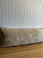 Front side of a long rectangular pillow with 'we are thankful' embroidered in white cursive on beige fabric.