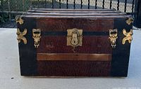 Front view of the antique wooden steamer trunk showing wood panels with dark reddish-brown grain, black trim, and brass-colored hardware including three locking clasps and a keyhole lock.