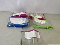 Multiple stacks of clear plastic pouches with colored zippers (pink, blue, red, green) arranged in groups on a table, showing quantity and color variety.