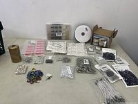 Overview of assorted craft and jewelry supplies laid on table, showing various small parts in plastic bags and containers, beads, and spools of ribbons.