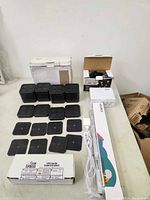 Photo showing multiple black rectangular pads arranged in two rows, several boxes, and a white electrical cord alongside a long narrow white box with color label.