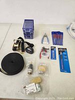 Wide view of miscellaneous tools on white surface including ratchet strap, pliers, blades, pens, and small hardware components.