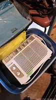 Image showing the white LIVING brand AC window fan placed inside a plastic storage bin, surrounded by other items.