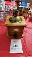 Front view of an antique wooden Tre Spade coffee grinder with brass top and handle, on a red tablecloth, including a small drawer with black knob and brand logo.