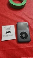 Front view of black Apple iPod Classic with screen and click wheel beside auction lot card and green tape on red fabric.