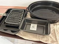 A group photo showing various baking and cooking items including a large black oval Dutch oven with lid, a muffin tin with six cups, a rectangular baking loaf pan with auction sticker, a square baking pan, and a metal grill rack with long handle.